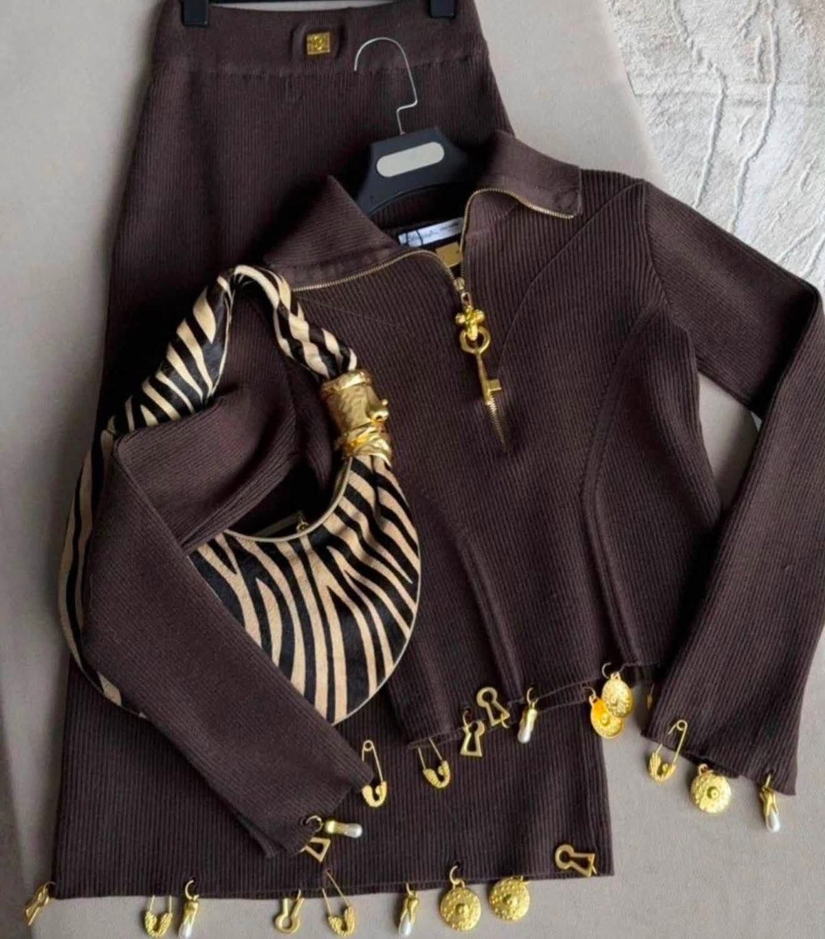 A dark brown ribbed knit three-piece outfit set consisting of a long-sleeve polo-style top with gold key and safety-pin embellishments, matching high-waisted skirt, and a zebra-print scarf, displayed on a hanger against a neutral background.