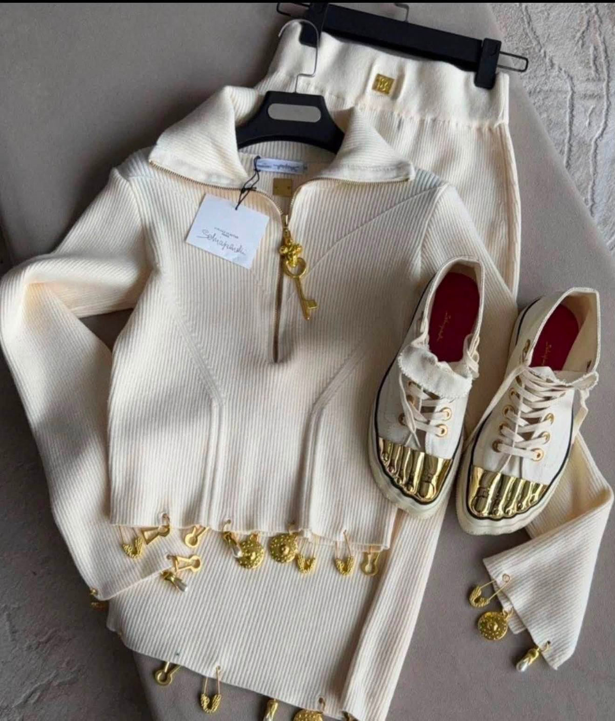 A cream ribbed knit zip-up hoodie with gold-tone hardware and dangling gold charms, shown with matching cream pants on hangers and a pair of cream high-top sneakers with metallic gold toe caps, laid out on a gray surface.