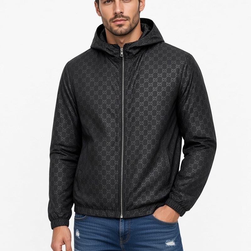 A man wearing a black hooded zip-up jacket with a subtle repeating monogram pattern, paired with blue jeans, against a plain white background.