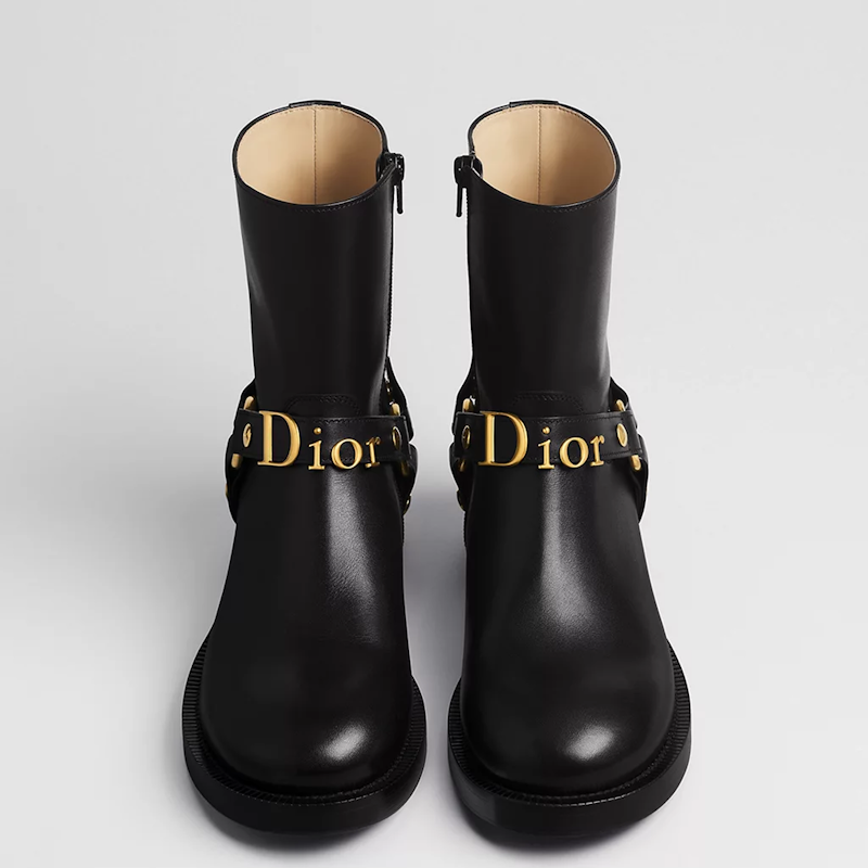 A pair of black leather ankle boots with gold-tone hardware and a prominent  logo strap, photographed from a front-facing angle on a light gray background.