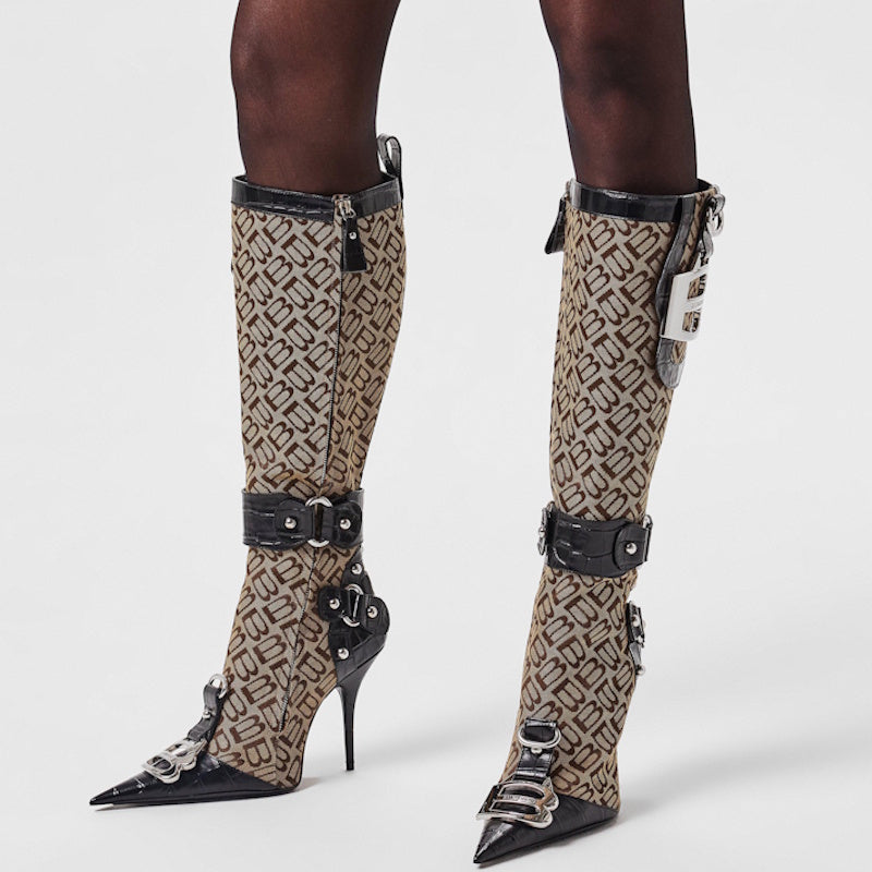 A pair of knee-high pointed-toe stiletto boots with a beige and black monogram pattern, black faux-leather straps, and silver hardware, shown on a model's legs against a light gray background.